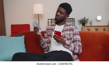 Planning Family Budget. Sad Poor African American Man Counting Money Cash, Use Mobile Phone Calculate Domestic Bills At Home. Worried Guy Upset Of Income And Saves Money For Planned Vacation, Gifts