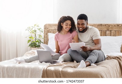 Planning Family Budget. Young Happy Black Couple Sitting On Bed At Home And Checking Financial Reports, Empty Space