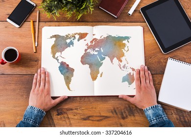 Planning His Journey. Top View Close-up Image Of Man Holding Hands On His Notebook With Colorful Map On It While Sitting At The Wooden Desk