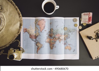 Planning The Trip. World Map With The Hat, Film Camera, Some Money, Notebook From Recycled Paper And Freshly Brewed Coffee Cup On The Dark Wooden Table Background.