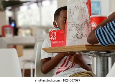 Plantation, FL / USA - 9/22/2020: African American Young Boy Child Seated At A Table In The Mall Food Court Being Good Eating A Chicken Sandwich, Fries And A Soft Drink With His Family And Bag Near.