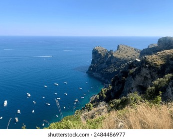 Plant-covered cliffside overlooking Southern Italian coastal cove with boats in the sea or ocean with a clear blue horizon. - Powered by Shutterstock