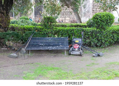 Plasencia,Spain - February 17, 2021:Municipal Gardener Worker Team Work Tools Leaning On Park Bench While Resting From Lawn Care. Professional Garden Equipment.