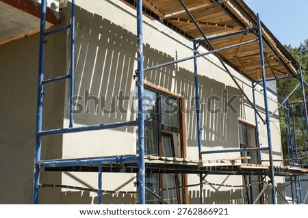 Plaster coating on facade insulation, external wall scaffolded for finishing, construction phase, smoothing render, surface preparation, house renovation works.