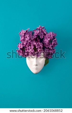 Plaster face, figurine decorated with lilac flowers. Lilac as a hairstyle on the head figurine against blue lasure background. Flat lay floral composition.