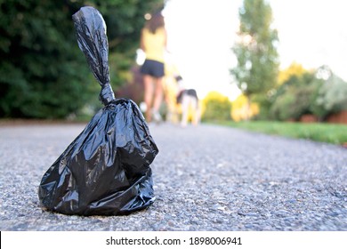 A plastic bag filled with dog poop left on sidewalk with woman and dog in the background.