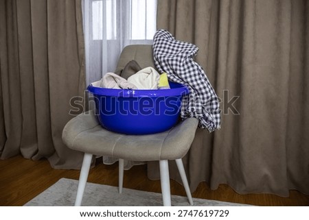 A plastic basin filled with laundry placed on a grey chair against neutral curtains.