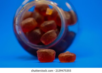 Plastic Bottle Full Of Gummy Vitamins Sideways With Gummies Falling Out In Front Of Blue Background.