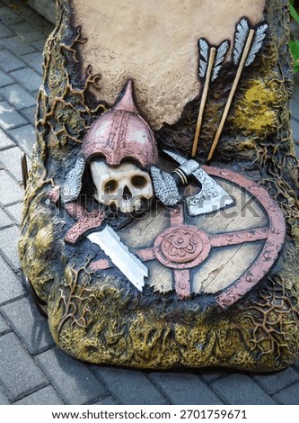 A plastic composition in the form of a skull, sword and axe on a shield with arrows on a bright sunny day. Playgrounds, sports, health, entertainment.