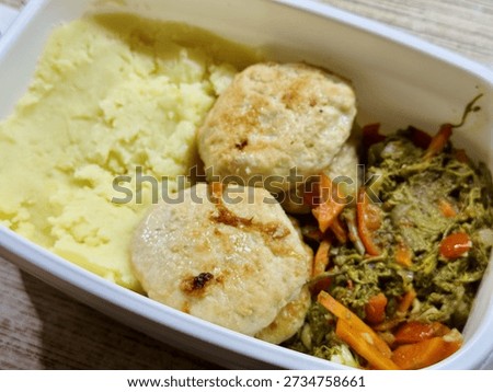 Plastic Container With Mashed Potatoes, Two PanFried Cutlets And SautEd Carrots And Greens On Wooden Desk, Midday Office Setting With Natural Light, BudgetFriendly Homemade Reheatable