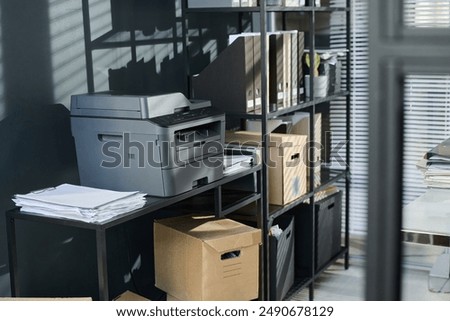 Plastic copier machine standing on desk with stack of paper documents by stand with shelves with cardboard boxes and folders with bills