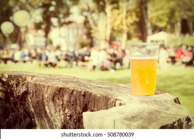 Plastic cup of beer on a tree trunk. In the background, blurred people participating in a village feast. Copy space. Vintage style photo.