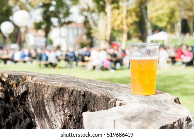 Plastic cup of beer on a tree trunk. In the background, blurred people participating in a village feast. Copy space