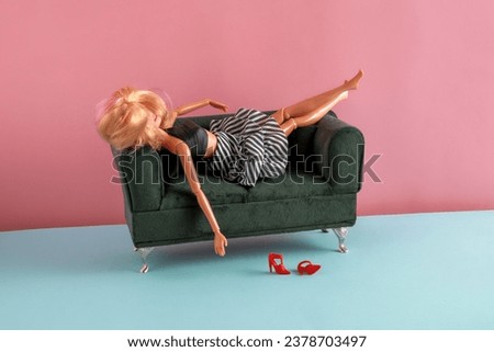 Plastic doll is lying on the sofa.  Pink and blue background. 