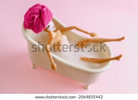 Plastic doll with towel on hair and facial moisturizing mask on face lying in bathroom. Spa day concept on pink background.