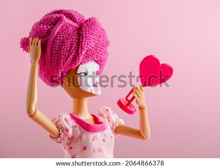 Plastic doll with towel on head and mask on face watching into mirror on a pink background. Spa and beauty care concept.