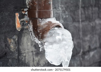 A Plastic Downspout, Downpipe Of A Roof Gutter System With Ice And Frozen Water In The Pipe Near A House Foundation In Winter.