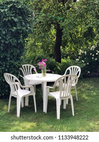 Plastic Furniture Set From Nineties In Super Green Garden Environment. White Chair And Table And Wild Flowers In The Glass. Vintage Feeling