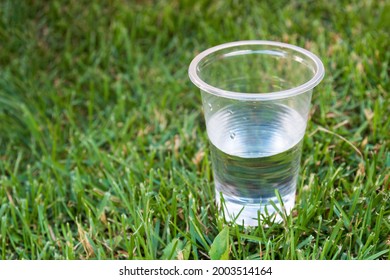 Plastic Glass With Water On Green Grass. Concept Background With Copy Space. Glass Half Empty Or Half Full