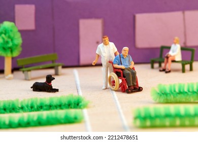 Plastic Miniature Male Nurse In Uniform Helping Senior Patient On Wheelchair In Front Of Hospice Building