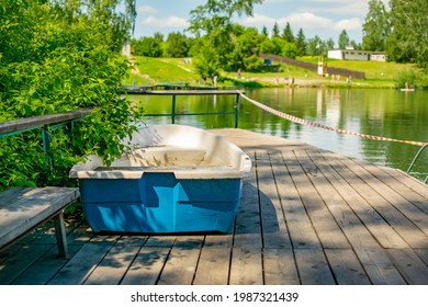 Plastic Old Broken With A Crack In The Hull Tourist Lake Boat Stands On A Wooden Pier Near The Reservoir On The Background Of The Beach With Vacationers Bathing People, Tourists