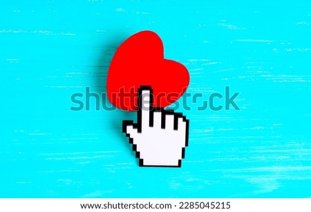 Plastic pointer cursor touches a red heart shape isolated on vibrant blue background. Digital technology and graphic design related concept.