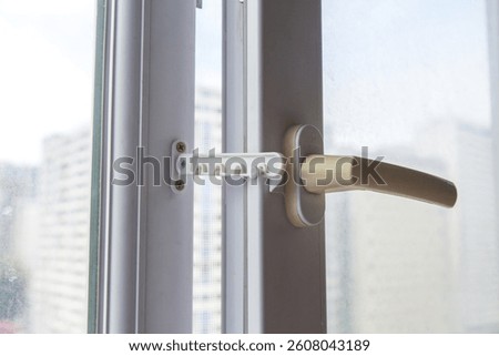 Plastic safety window limiter with beige handle installed on white frame, partially open window with blurred city view, ensuring ventilation and child safety, concept of home protection