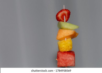 A Plastic Skewer Has Cut Pieces Of Juicy Fruit: Strawberry, Honeydew Melon, Cantaloupe, Pineapple And Watermelon, And Is Held Up Before A White Background.