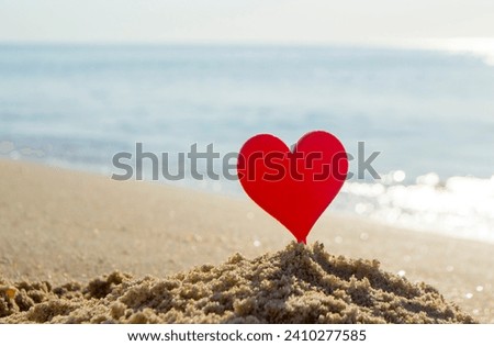 Plastic stick in shape of red heart in sand on sandy beach of sea shore on background of sea waves on sunny summer day close-up. Concept Love relationship romance amorousness amour St. Valentines Day