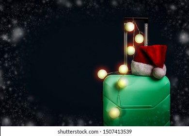 Plastic Suitcase, Santa Claus Cap And Garland On A Dark Blue Background With Snow. Concept Of Travel, Business Trips, Trips To Visit Friends And Relatives On Christmas Holidays. New Year's Journey