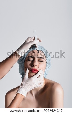 plastic surgeon in latex gloves holding scalpel near patient in surgical cap with marked lines on face isolated on grey