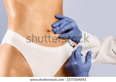 Plastic surgeon in protective gloves making injection at belly area for unrecognizable lady, blue studio background. Slim woman having lipolysis treatment at beauty salon, cropped close up view