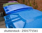 Plastic Waste Recycling Wheelie Bins lined up and secured in a UK garden storage area beside house brick wall, waiting for weekly council collection close-up