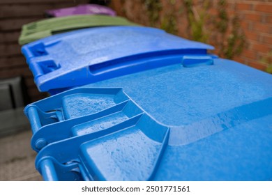 Plastic Waste Recycling Wheelie Bins lined up and secured in a UK garden storage area beside house brick wall, waiting for weekly council collection close-up