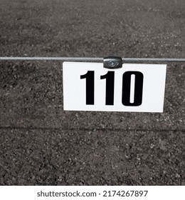 A Plastic White Plate With The Number Of A Free Parking Space Hangs On An Iron Cable Against The Background Of Asphalt During The Day.