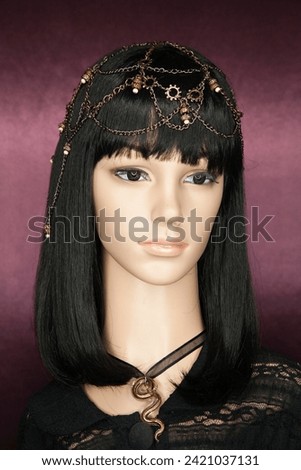 Plastic woman mannequin with long black hair wearing a chain head piece with beads, and a black lace on a purple background. Ring light catchlights in her eyes.