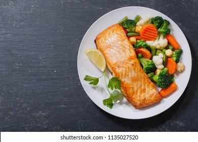 Plate Of Baked Salmon Steak With Vegetables On Dark Background, Top View