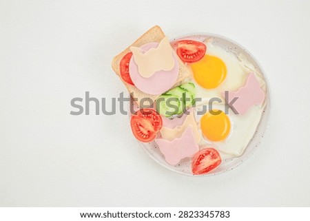 Plate of breakfast items including fried eggs, sliced tomatoes, cucumber, and toast with butterfly-shaped ham and cheese, presented on a white background