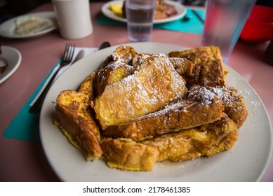 A plate of challah French toast on a white plate in a diner with a pink and blue table. The breakfast food has toppings of powdered sugar and real maple syrup. - Shutterstock ID 2178461385