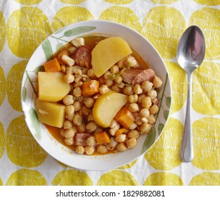Plate of chickpeas with potatoes, carrots, ham and sausage over a yellow tablecoth with a steel spoon. - Shutterstock ID 1829882081