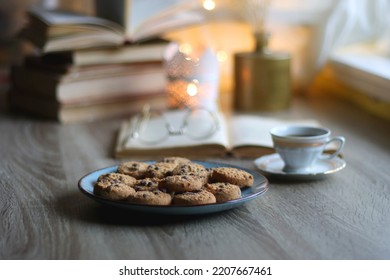 Plate of chocolate chip cookies, stack of vintage books, reading glasses, cup of tea or coffee, lit candle and fairy lights. Hygge at home. Selective focus. - Powered by Shutterstock