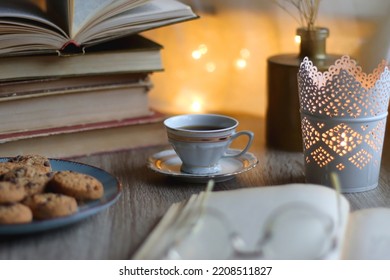 Plate of chocolate chip cookies, stack of vintage books, reading glasses, cup of tea or coffee, lit candle and fairy lights. Hygge at home. Selective focus. - Powered by Shutterstock