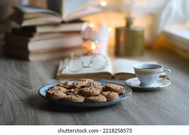 Plate of chocolate chip cookies, stack of vintage books, reading glasses, cup of tea or coffee, lit candle and fairy lights. Hygge at home. Selective focus.