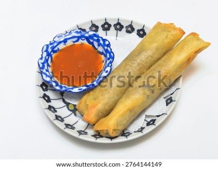 A plate of crispy spring rolls accompanied by a small bowl of vibrant red dipping sauce, all on a white plate with a blue and black floral pattern