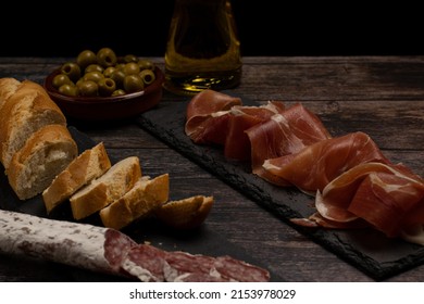 plate of cured Iberian Serrano ham, cured meats on a slate stone plate, on a wooden table with olives, garlic, olive oil and bread. 