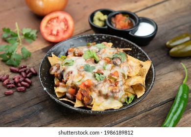 A Plate Of Delicious Corn Chips Nachos In Cheddar Cheese With Beans, Tomato, Green Chili, Black Olives, Cilantro Served With Guacamole, Sour Cream And Tomato Salsa Dip On The Side Isolated On The Wood