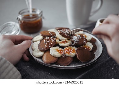 A Plate Filled With Chocolate Mini Pancakes And Banana Slices Topped With Salted Caramel. Hands Holding A Fork On Which A Pancake.