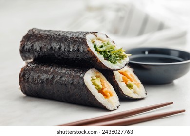 A plate filled with a variety of sushi rolls with a set of wooden chopsticks and bowl of soy sauce, placed on a simple kitchen table. - Powered by Shutterstock