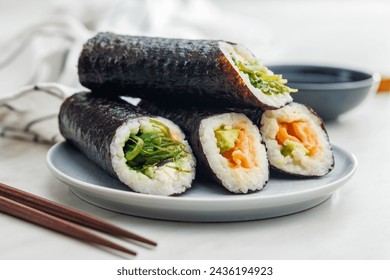 A plate filled with a variety of sushi rolls with a set of wooden chopsticks and bowl of soy sauce, placed on a simple kitchen table. - Powered by Shutterstock