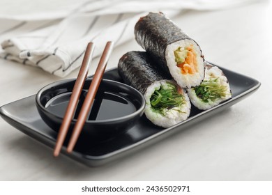 A plate filled with a variety of sushi rolls with a set of wooden chopsticks and bowl of soy sauce, placed on a simple kitchen table. - Powered by Shutterstock